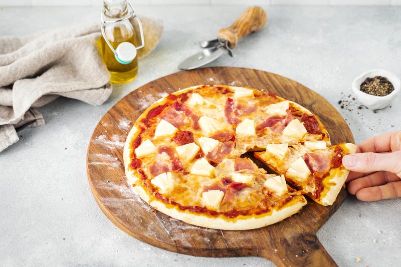 Hawaiipizza, ein Stück wird mit einer Hand angehoben.