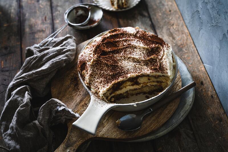 Tiramisu in einer weißen Schale auf einem Holzbrett, daneben ein graues Tuch